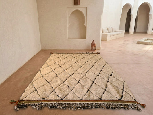 A large rectangular Moroccan-style rug with a raised diamond pattern and fringe tassels, displayed on a terracotta floor in a minimalist, sunlit room with arched alcoves.