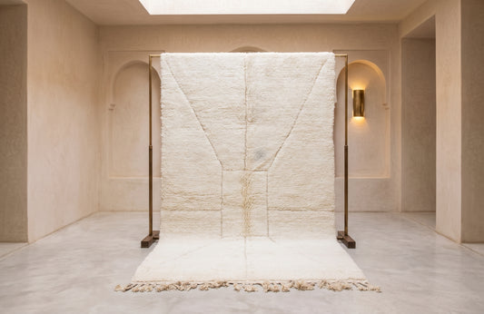 A large off-white textured rug displayed on a black metal stand in a minimalist, warm-toned interior room with beige walls and a light marble floor.