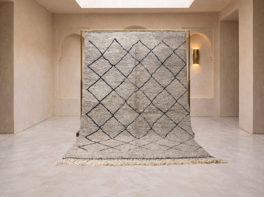 A large woven area rug with a black diamond lattice pattern on a light beige and gray textured background, hanging from a wooden frame in a minimalist beige room.