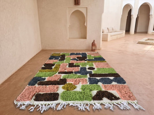 A rectangular shag area rug with a multicolor geometric patchwork pattern and white fringe, displayed on a terracotta tiled floor in a Moroccan-style courtyard with arched alcoves.