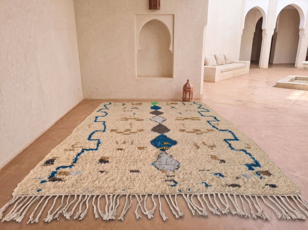 A rectangular woven area rug with fringe, displayed in a Moroccan-style room with beige walls, arched alcoves, and a terracotta tile floor. The rug has a cream base with blue, gray, and brown geometric patterns.