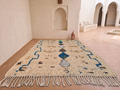A rectangular woven area rug with fringe, displayed in a Moroccan-style room with beige walls, arched alcoves, and a terracotta tile floor. The rug has a cream base with blue, gray, and brown geometric patterns.