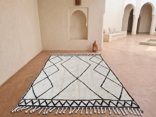 A rectangular area rug with a black geometric diamond pattern on an off-white background, laid out on a terracotta-colored floor in a Moroccan-style courtyard with arches and a bench in the background.