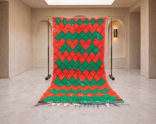 A large Moroccan-style rug with a vibrant red and green harlequin diamond pattern, displayed hanging from a metal stand in a minimalist beige gallery space.