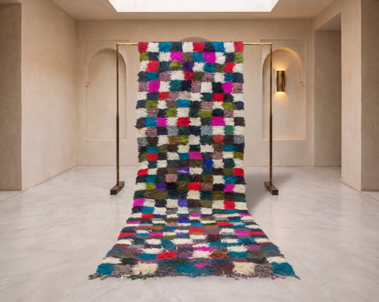 A multicolor shaggy patchwork rug displayed on a metal stand in a minimalist beige gallery space.