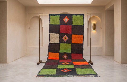 A large patchwork wool rug with geometric diamond motifs hangs from a metal stand in a minimalist, warm-toned gallery space with beige walls and arches.