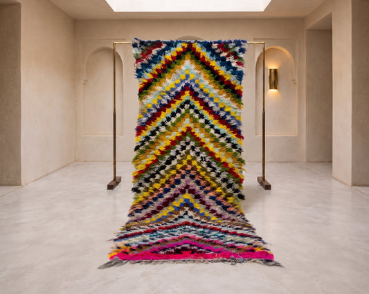 A large, vibrant, multicolored woven rug with a textured chevron pattern, displayed hanging from a metal stand in a minimalist, warm-toned gallery space.
