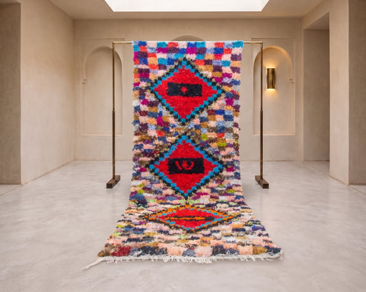A colorful Moroccan-style woven rug displayed on a metal stand in a minimalist, warm-toned gallery space with arched walls and a light marble floor.