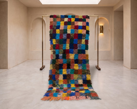 A large, multicolored textured rug with a patchwork grid pattern hangs from a minimalist metal stand in a modern, warm-toned gallery space with beige walls and arched alcoves.