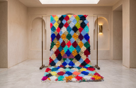 A vibrant, multicolored shag rug with a diamond pattern, displayed on a black metal stand in a minimalist beige room with arched walls and a matching rug on the floor.
