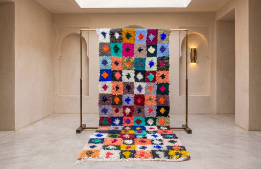 A colorful Moroccan-style shag rug displayed on a metal stand in a minimalist beige room, with a matching flat view of the rug laid out in front.