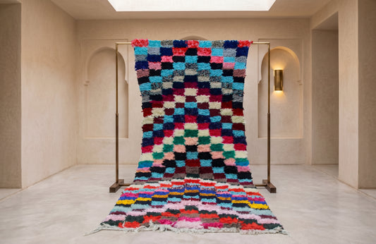 A colorful Moroccan-style shag wool rug with a geometric checkerboard pattern hangs from a metal display stand in a minimalist, warm-toned gallery space.