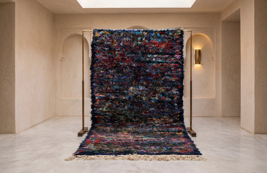 A large, textured shag rug with a multicolor abstract pattern in blue, red, black, and white, hanging from a wooden frame in a minimalist beige gallery space.