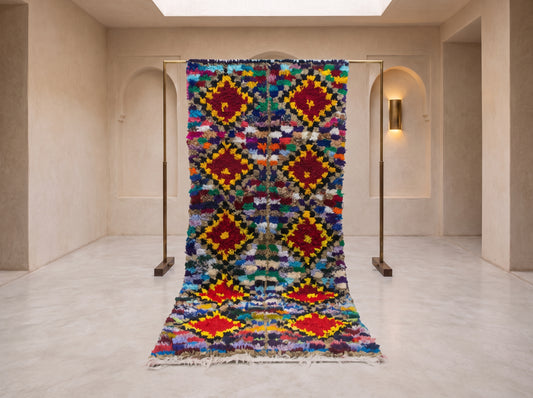 A large, vibrant Moroccan-style woven rug displayed on a minimalist brass stand in a modern, neutral-toned gallery space.