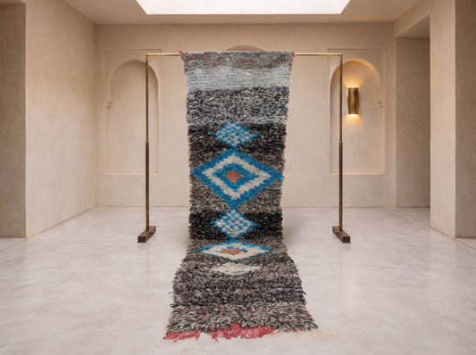 A long shag area rug with a geometric diamond pattern in blue, gray, black, and red, hanging from a brass rack in a minimalist beige room.