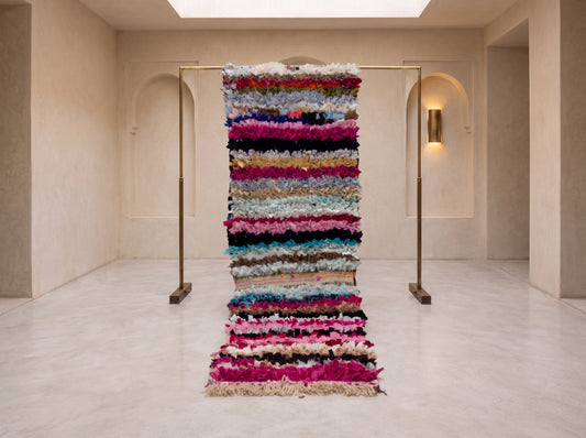 A tall, narrow multicolored shag rug displayed on a minimalist indoor gallery floor, suspended between two slim brass stands in a cream-colored room with arched wall niches.