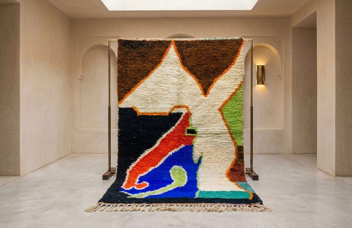 A large abstract area rug displayed on a metal stand in a minimalist beige gallery space. The rug features a bold, geometric pattern with large shapes in brown, cream, black, blue, red, green, and orange, finished with a fringed edge.