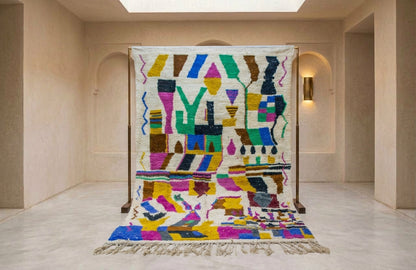 A large woven area rug with a colorful geometric pattern is displayed on a wooden stand in a minimalist beige gallery space.