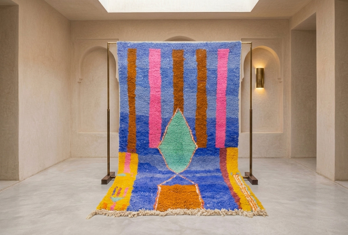 A colorful Moroccan-style woven rug displayed on a metal stand in a minimalist, warm-toned gallery space. The rug features a vibrant blue base with vertical pink and brown stripes, a central mint green teardrop motif, and yellow accents near the fringe.