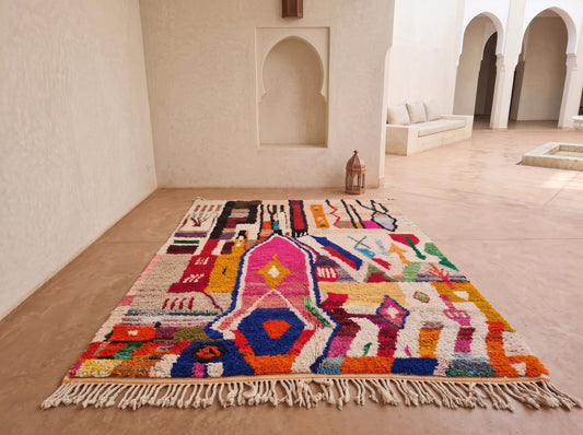 A vibrant Moroccan-style area rug with a colorful geometric and tribal pattern, laid out on a sandy floor in a minimalist, sunlit room with arched alcoves.