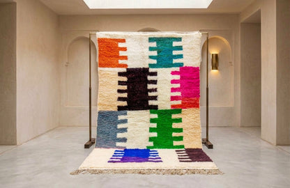 A multicolor geometric patterned wool rug displayed on a black metal stand in a minimalist beige room with arched doorways.