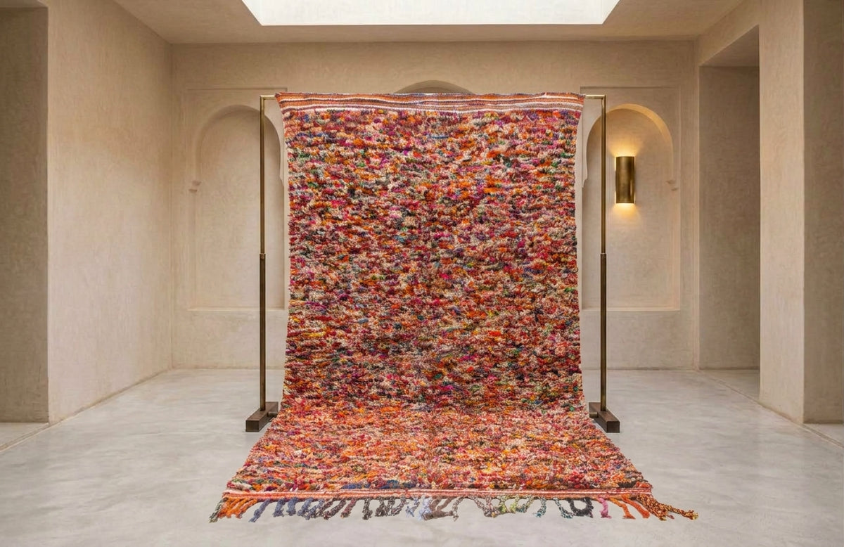 A large, vibrant Moroccan-style woven rug displayed on a black metal stand in a minimalist, warm-toned gallery space.