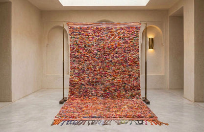 A large, vibrant Moroccan-style woven rug displayed on a black metal stand in a minimalist, warm-toned gallery space.