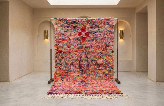 A large, vibrant, multicolored shag rug displayed on a black metal stand in a minimalist, warm-toned interior room with beige walls and arched alcoves.