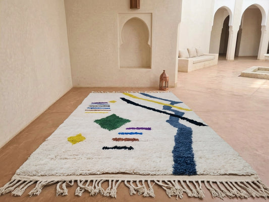 A rectangular white shag rug with colorful abstract geometric patterns and a fringed edge, displayed on a terracotta floor in a minimalist Moroccan-style room with arched alcoves.