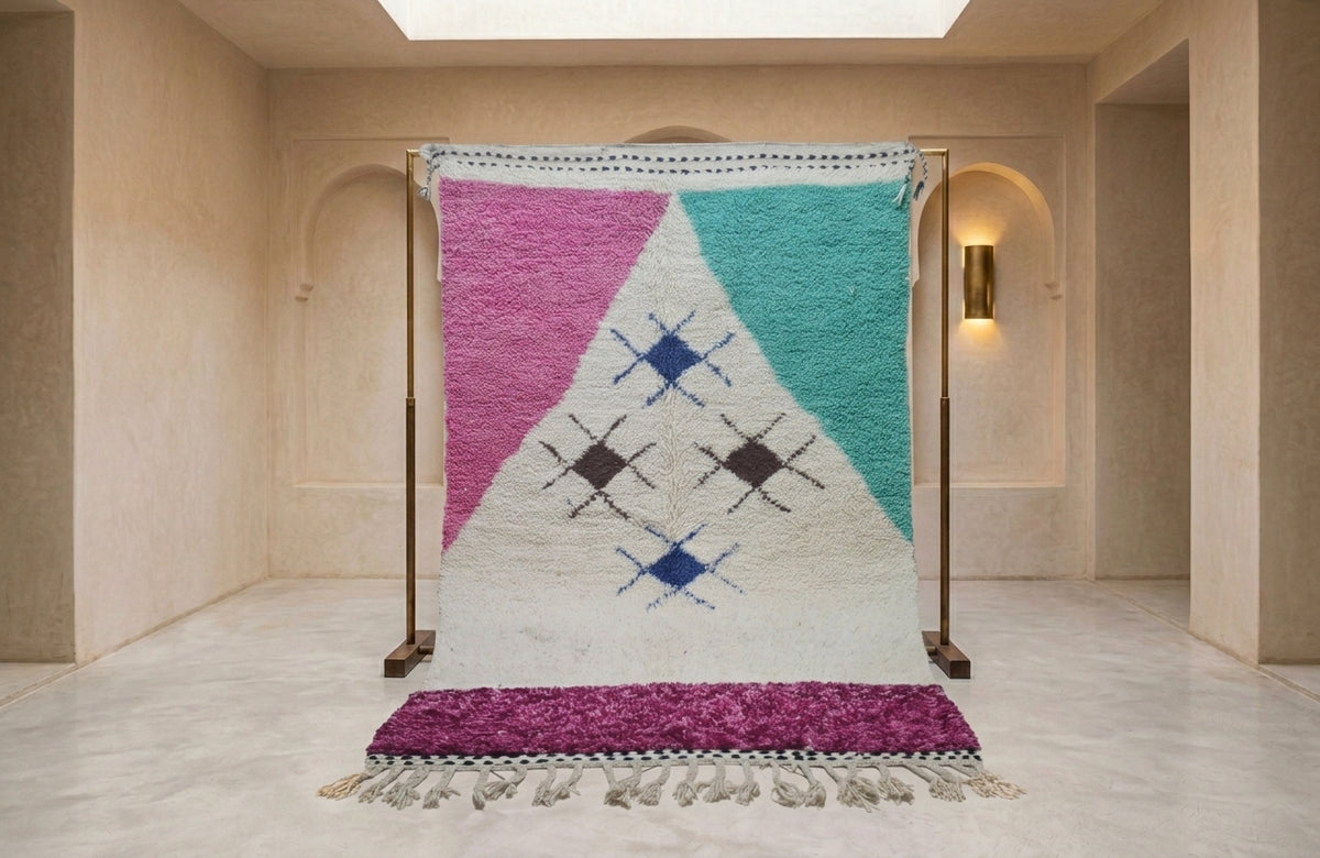 A large woven Moroccan-style rug displayed on a wooden stand in a minimalist beige hallway. The rug features a geometric pattern with pink, teal, and white sections, accented with blue and black diamond motifs, and finished with a purple fringe.