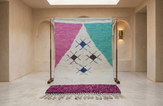 A large woven Moroccan-style rug displayed on a wooden stand in a minimalist beige hallway. The rug features a geometric pattern with pink, teal, and white sections, accented with blue and black diamond motifs, and finished with a purple fringe.