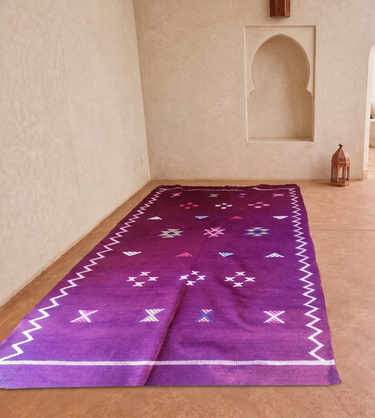 A purple Moroccan-style wool rug with white geometric patterns and borders, laid out on a terracotta tiled courtyard floor in front of cream-colored stucco walls with arches.