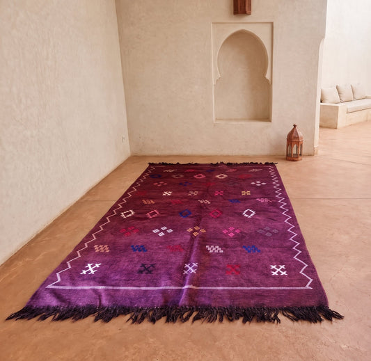 A rectangular purple Moroccan-style rug with black fringe and geometric patterns, displayed on a terracotta-colored floor in a traditional courtyard with white stucco walls and arches.