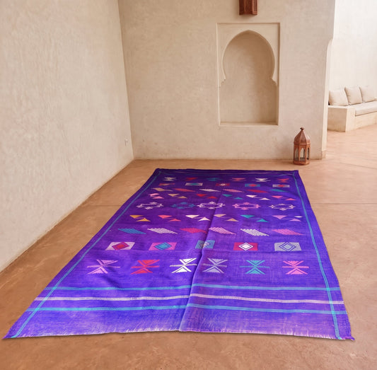 A rectangular purple woven rug with colorful geometric patterns and fringe, laid out on a terracotta tiled floor in a Moroccan-style courtyard with beige walls and arches.