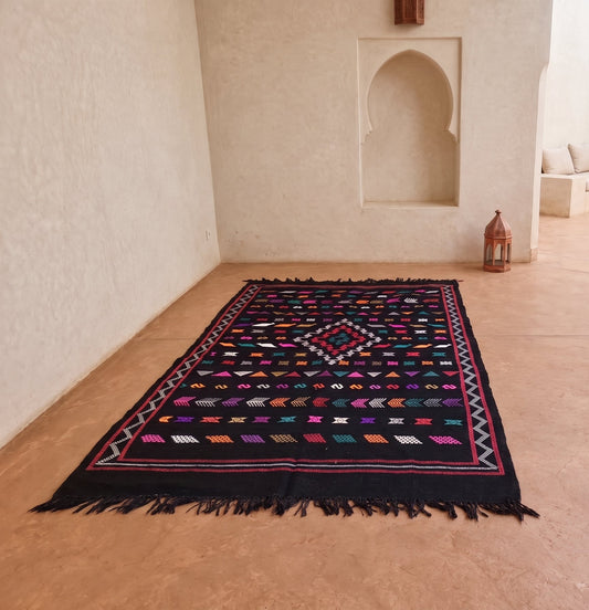 A rectangular black Moroccan-style rug with colorful geometric patterns and fringe, displayed on a terracotta tiled floor in a minimalist, cream-colored courtyard.