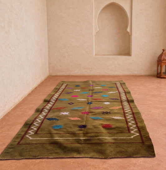 A rectangular olive green area rug with colorful geometric patterns and a white zigzag border, displayed on a terracotta-colored floor in a Moroccan-style room with arched wall niches.