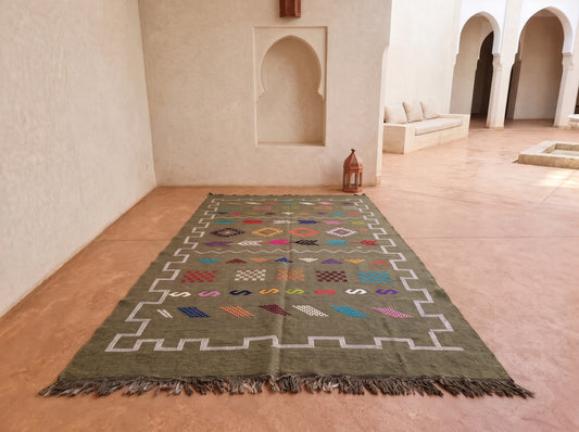 A rectangular Moroccan-style wool rug with a muted olive green base, white geometric border, and multicolored diamond and zigzag motifs, displayed on a terracotta tiled floor in a Moroccan courtyard.