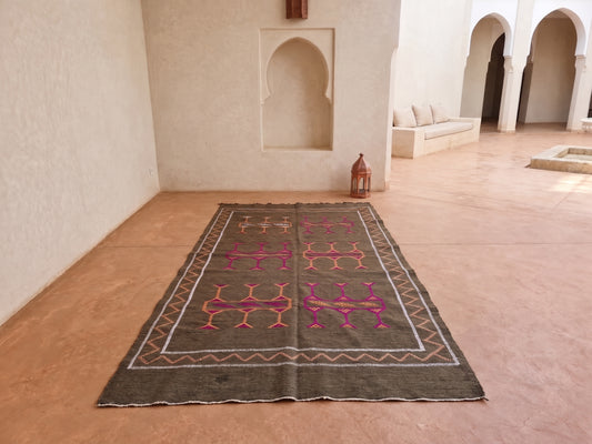 A rectangular Moroccan-style area rug with a dark brown base and pink geometric patterns, displayed on a terracotta tiled floor in a Moroccan courtyard with white stucco walls and arches.