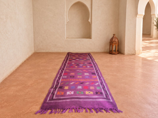 A purple Moroccan-style prayer rug with colorful geometric patterns and fringed edges, laid out on a terracotta tiled floor in a sunlit room with arched wall niches.