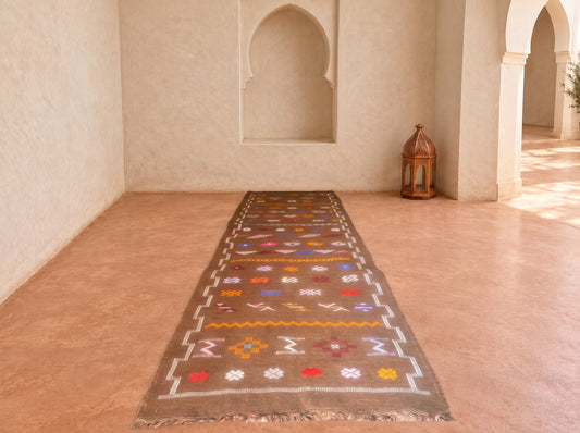 A long rectangular runner rug with a brown base and colorful geometric patterns, laid out on a terracotta-colored floor in a Moroccan-style room with arches and a lantern.