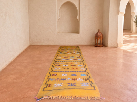A long yellow Moroccan-style runner rug with colorful geometric patterns and fringe, displayed on a terracotta floor in a Moroccan-style room with arches and a lantern.
