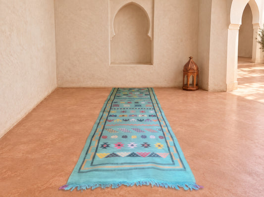A turquoise blue patterned runner rug with fringed ends is displayed on a terracotta-colored floor in a Moroccan-style room with arched wall niches and a wooden lantern.