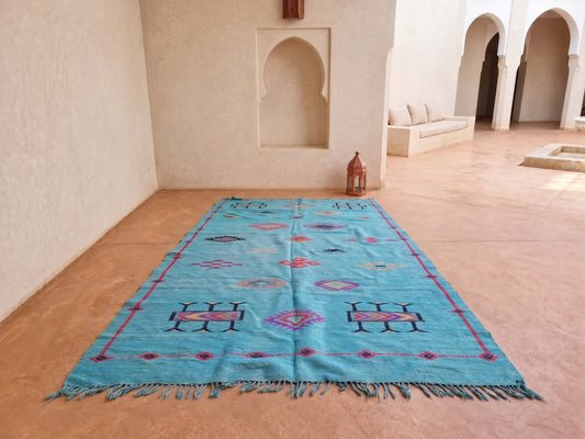 A rectangular turquoise blue Moroccan-style rug with geometric patterns and fringe, laid out on a terracotta tiled floor in a sunlit courtyard with white stucco walls and arches.