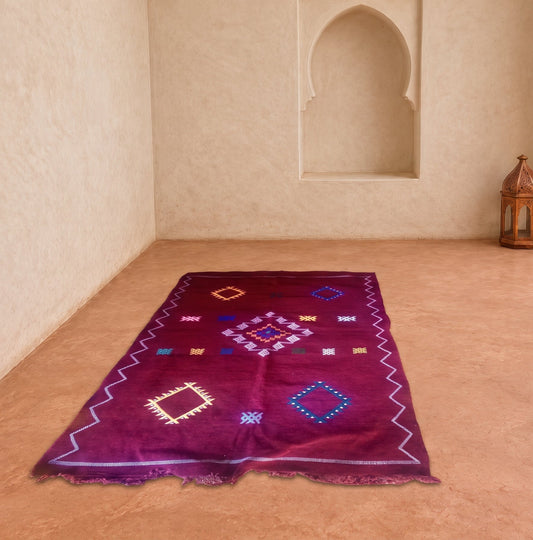 A rectangular Moroccan-style wool rug in deep magenta purple, laid out on a terracotta tiled floor in a sunlit room with beige stucco walls and arches.