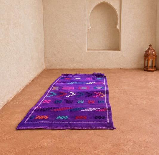A purple Moroccan-style woven rug with colorful geometric patterns is laid out on a terracotta tiled floor in a sunlit room with beige walls and arches.