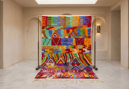 A vibrant, multicolored Moroccan-style woven rug displayed on a wooden stand in a minimalist, warm-toned interior room with beige walls and arches.