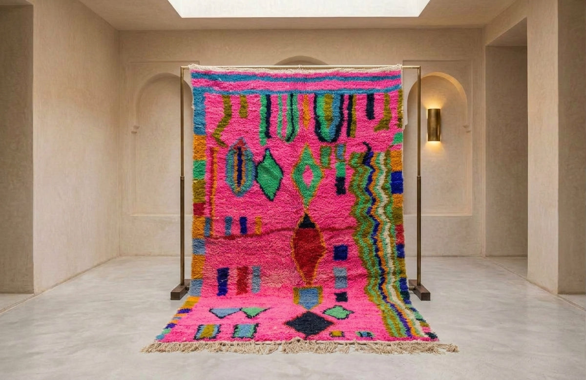 A vibrant Moroccan-style wool rug displayed on a metal stand in a minimalist beige hallway. The rug features a bright pink base with multicolored geometric patterns including green, blue, yellow, and red, and is finished with a fringed edge.