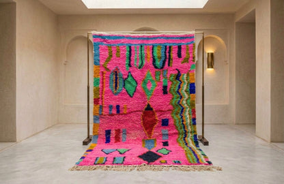 A vibrant Moroccan-style wool rug displayed on a metal stand in a minimalist beige hallway. The rug features a bright pink base with multicolored geometric patterns including green, blue, yellow, and red, and is finished with a fringed edge.