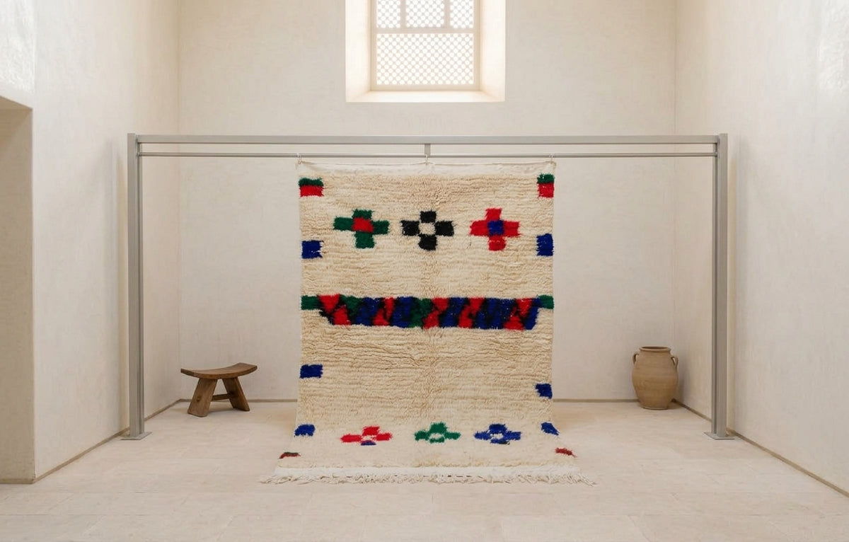 A textured, hand-woven Moroccan-style rug with a cream base and colorful geometric patterns, hanging from a metal frame in a minimalist, sunlit room.