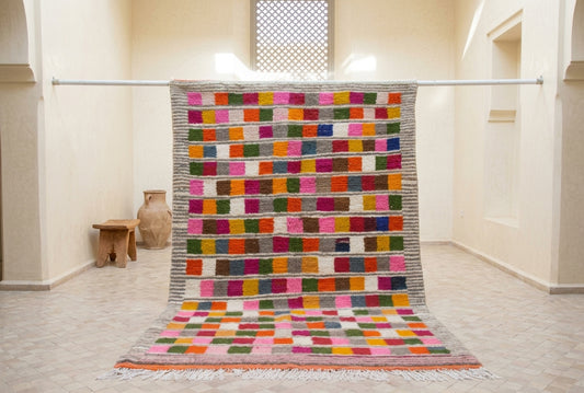 A large rectangular woven rug with a colorful grid pattern of square patches in pink, orange, green, blue, yellow, and red, hanging from a metal rod in a minimalist beige room.
